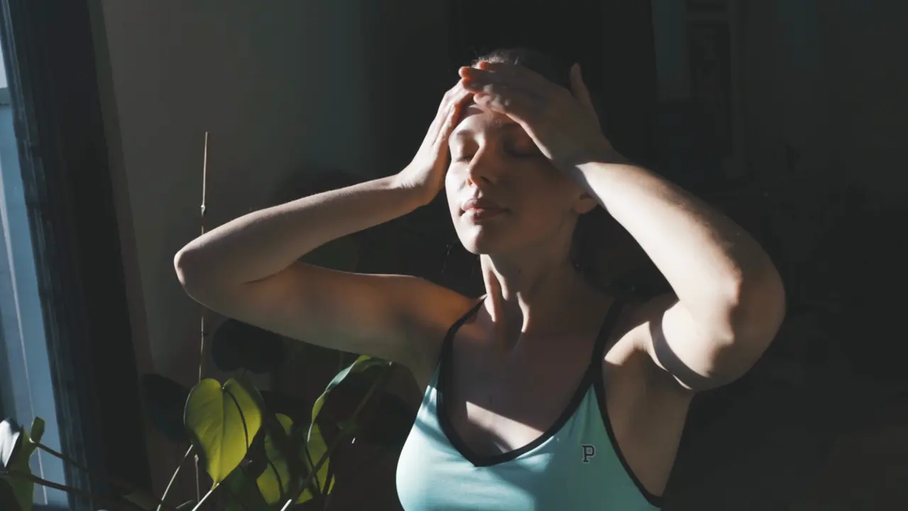 A woman by a sunny window pressing her hands to her forehead