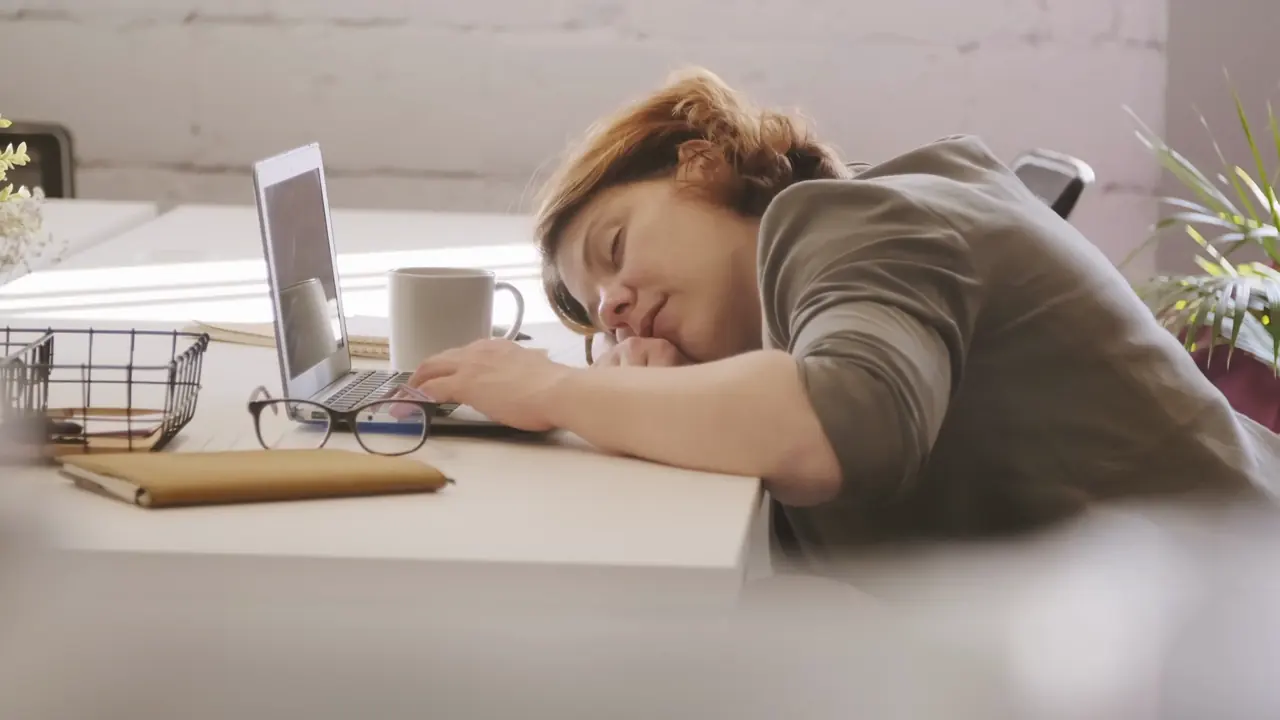 A woman asleep at her desk beside a laptop and a mug